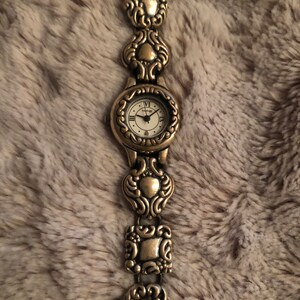 May include: A gold-toned wristwatch with a white face and Roman numeral hour markers. The watch band is made of linked, ornate, gold-toned metal pieces.