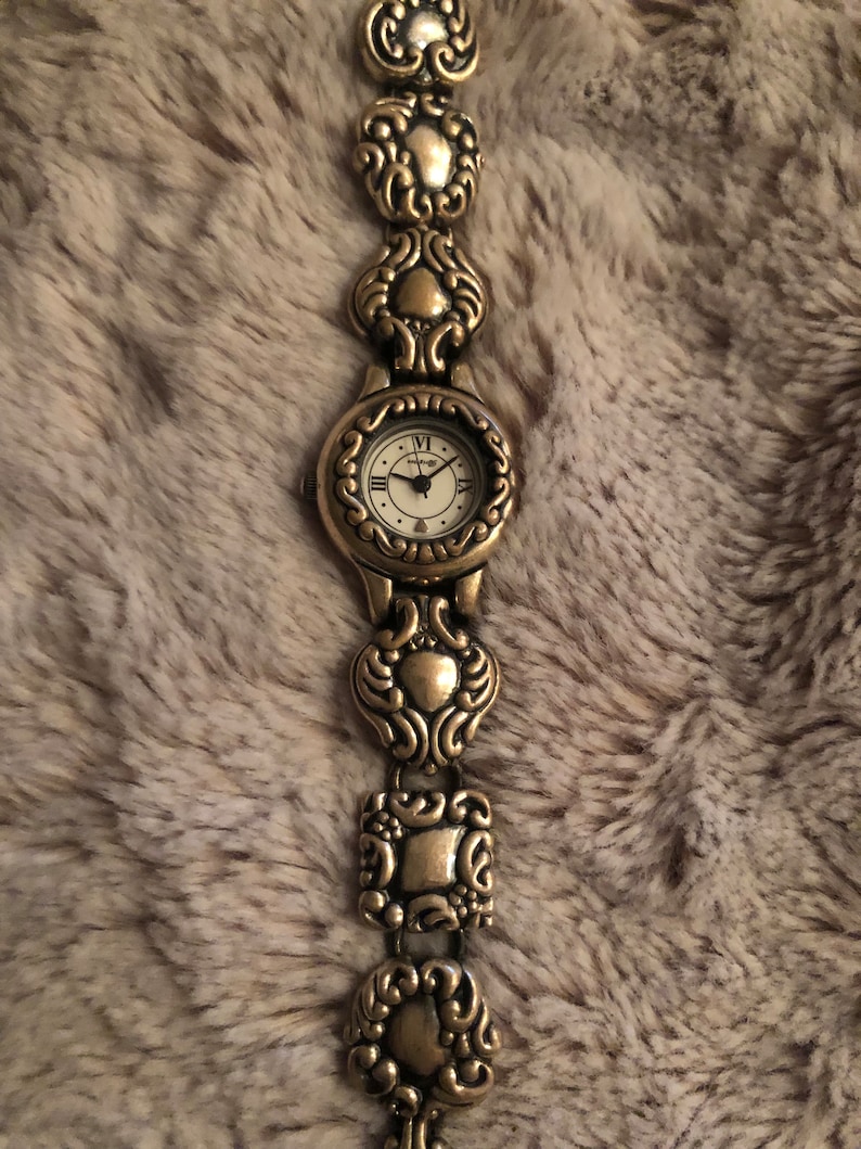 May include: A gold-toned wristwatch with a white face and Roman numeral hour markers. The watch band is made of linked, ornate, gold-toned metal pieces.