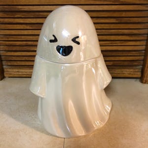 May include: A white ceramic ghost figurine with a smiling face. The ghost has a round head and a flowing body.
