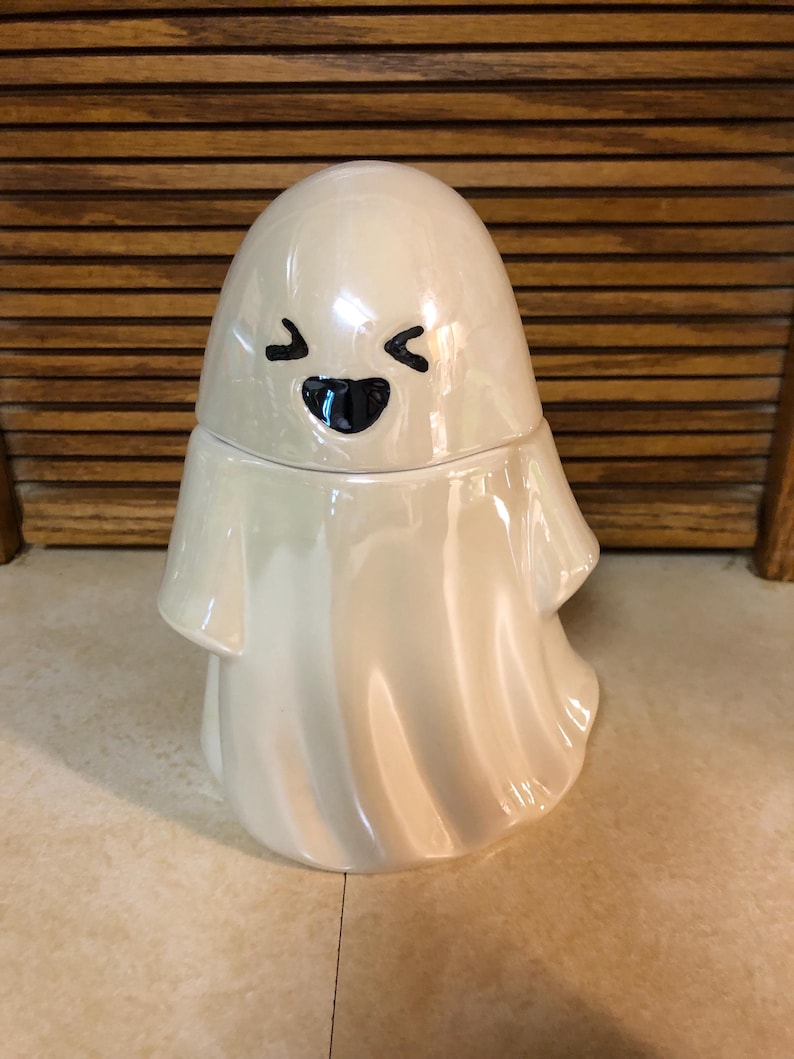 May include: A white ceramic ghost figurine with a smiling face. The ghost has a round head and a flowing body.