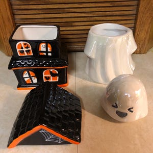 May include: A set of four Halloween-themed ceramic candle holders. The set includes a black and orange house with a roof, a white ghost with a smiling face, and two white ghosts with no facial features.