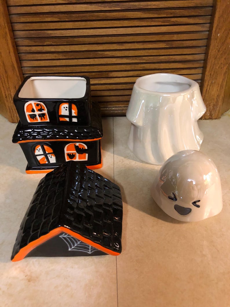 May include: A set of four Halloween-themed ceramic candle holders. The set includes a black and orange house with a roof, a white ghost with a smiling face, and two white ghosts with no facial features.