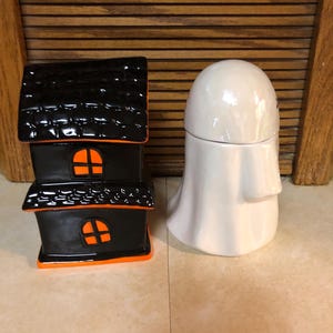May include: A black and orange ceramic house shaped like a haunted house and a white ceramic ghost. Both are Halloween themed decorations.