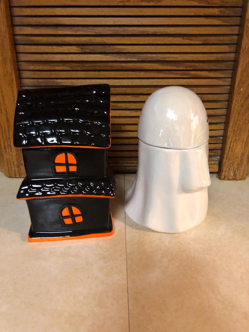 May include: A black and orange ceramic house shaped like a haunted house and a white ceramic ghost. Both are Halloween themed decorations.