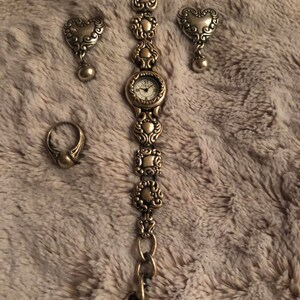 May include: A gold-toned watch with a heart-shaped design on the band. The watch comes with a pair of matching heart-shaped earrings and a ring.