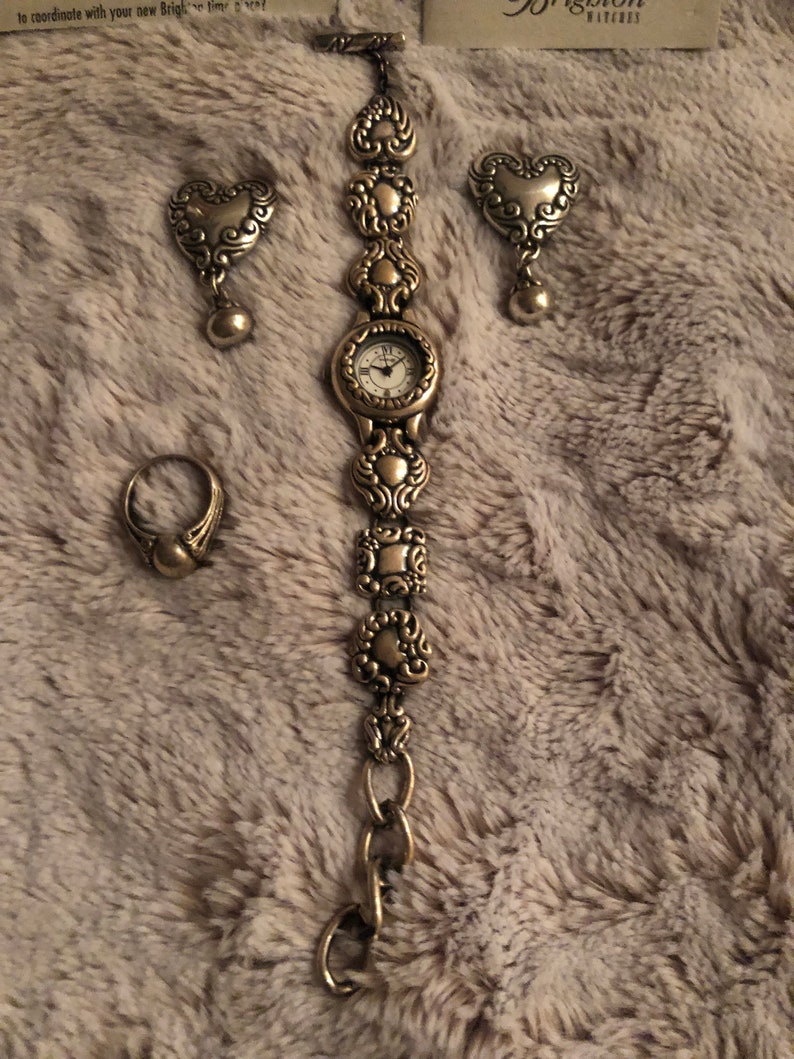 May include: A gold-toned watch with a heart-shaped design on the band. The watch comes with a pair of matching heart-shaped earrings and a ring.