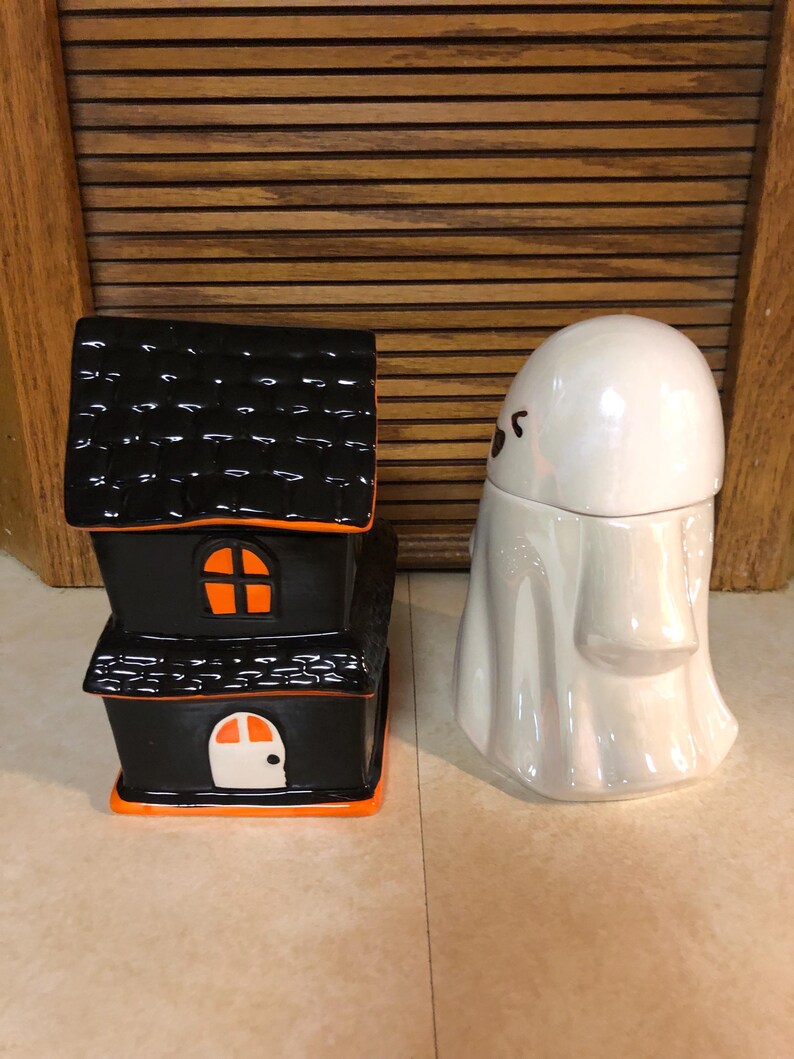 May include: A black and orange ceramic haunted house and a white ceramic ghost figurine. Both are Halloween themed decorations.