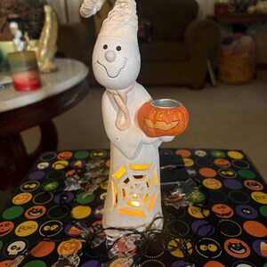 May include: A white ceramic ghost figurine wearing a tall, pointed hat, holding a small orange jack-o'-lantern candle holder. The ghost features a cutout spiderweb design on its body, illuminated from within by a warm light, surrounded by a string of clear ghost-shaped lights.