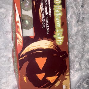 May include: A rectangular box containing 10 Halloween lights, featuring a graphic of a jack-o'-lantern. The packaging states indoor/outdoor use, a lighted length of 8.5ft, and a total length of 10ft. Two iridescent ghost-shaped light covers are visible.