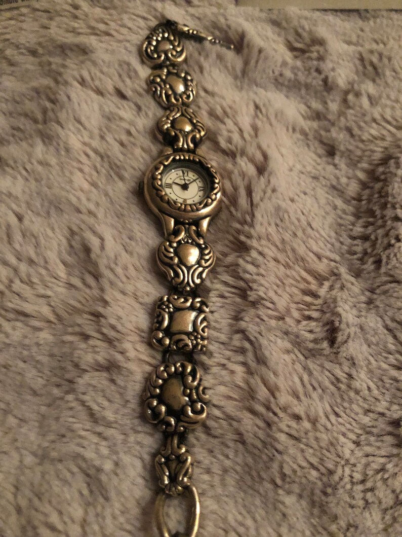 May include: A silver watch with a white face and ornate silver band. The band is made up of a series of linked, decorative, heart-shaped pieces.