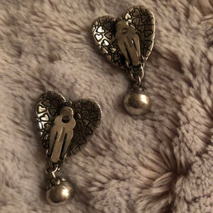 May include: A pair of silver heart-shaped earrings with a textured surface and a dangling ball charm.