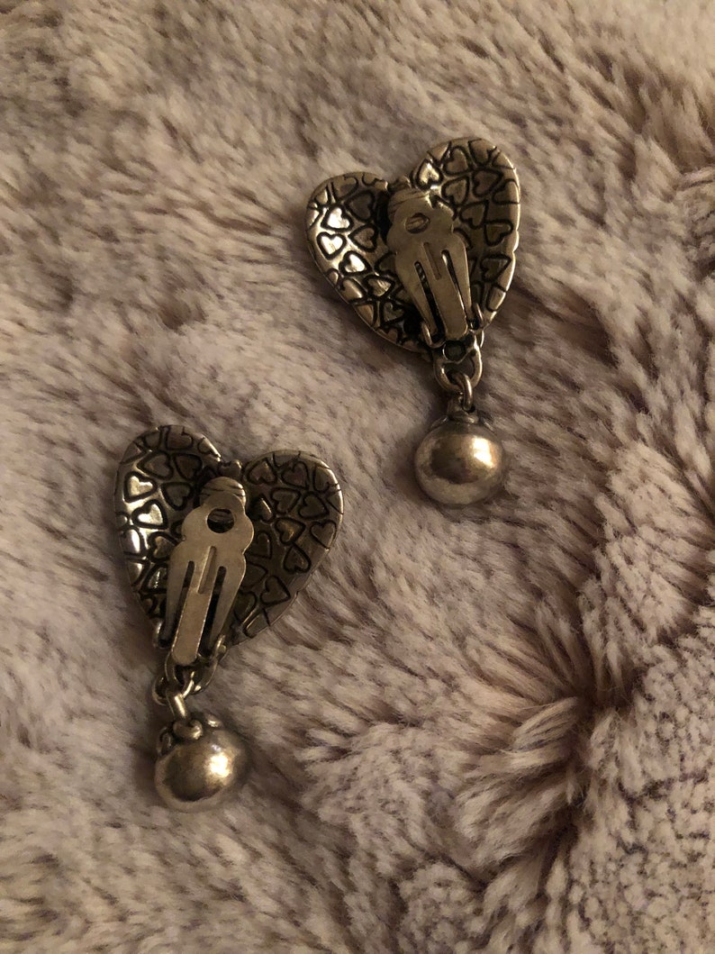 May include: A pair of silver heart-shaped earrings with a textured surface and a dangling ball charm.