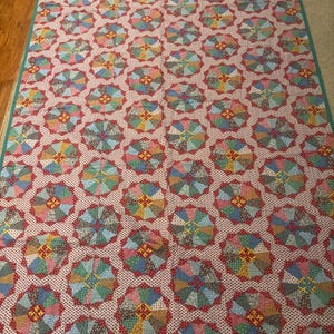 May include: A rectangular quilt featuring a repeating pattern of circular motifs composed of multi-colored fabric segments. The background fabric is white with small red dots, and the entire quilt is bordered by a solid teal fabric trim.