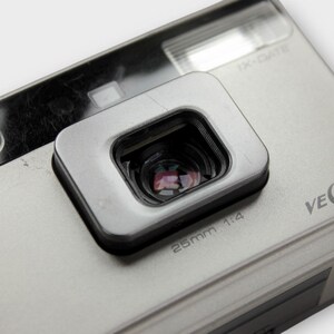 May include: Close-up of a silver Minolta Vectis UC camera. The camera has a rectangular viewfinder and a 25mm 1:4 lens. The camera body is silver with the model name printed on the side. The camera has a flash and a small button.