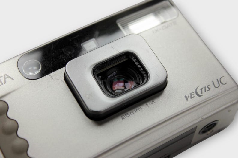 May include: Close-up of a silver Minolta Vectis UC camera. The camera has a rectangular viewfinder and a 25mm 1:4 lens. The camera body is silver with the model name printed on the side. The camera has a flash and a small button.