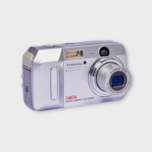 May include: Silver Olympus Camedia digital camera with a zoom lens. The camera has a 5.0 megapixel resolution and features the text "OLYMPUS" and "CAMEDIA DIGITAL CAMERA C-500 ZOOM". The lens has the text "OLYMPUS LENS" and "AF ZOOM 6.3-18.9mm 1:2.8-4.9".