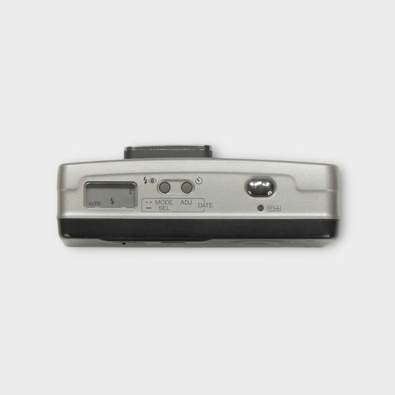 May include: Silver and black point-and-shoot camera. The camera has a small LCD screen and buttons labelled "AUTO", "MODE/SEL", "ADJ", and "DATE". The camera also has an "ON" button and a flash symbol.