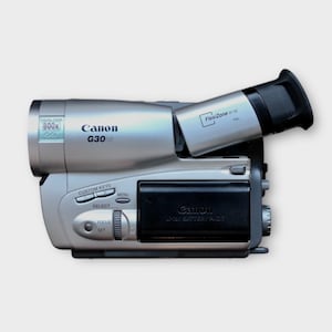 May include: A silver Canon G30 camcorder with a black lens and a black battery. The camcorder has a zoom lens, a built-in microphone, and a LCD screen.