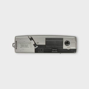 May include: A silver and black Minolta camera, seen from below. The camera features a battery compartment, a small port, and the text "76606250 MINOLTA CO., LTD OSAKA, JAPAN MADE IN INDONESIA".