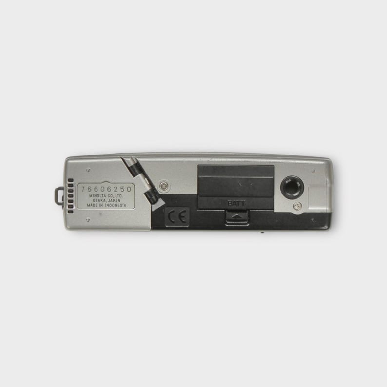 May include: A silver and black Minolta camera, seen from below. The camera features a battery compartment, a small port, and the text "76606250 MINOLTA CO., LTD OSAKA, JAPAN MADE IN INDONESIA".