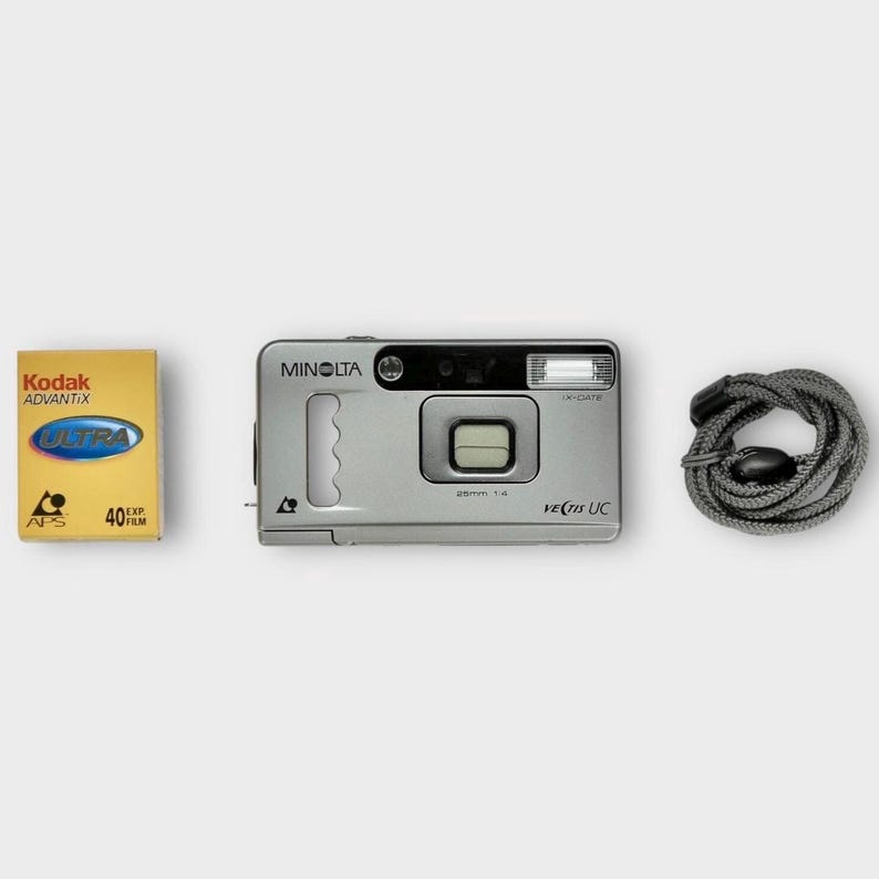 May include: A silver Minolta Vectis UC camera with a 25mm f/4 lens, a roll of Kodak Advantix Ultra 40 film, and a grey camera strap. The camera has a built-in flash and a rectangular viewfinder. The film is in a yellow box.