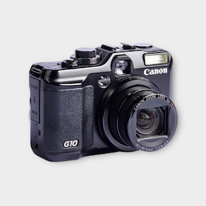 May include: Black Canon G10 digital camera with a zoom lens. The camera has a built-in flash and the Canon logo is visible. The lens is labeled "Canon Zoom Lens 5x". The camera has a textured grip and various buttons and dials.