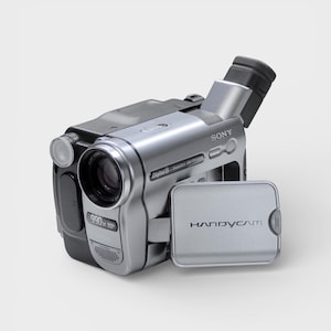 May include: Silver Sony Handycam camcorder with a black top and a flip-out screen. The camcorder has a lens, a viewfinder, and the text "990x" on the side. The word "SONY" is printed on the side of the camcorder.