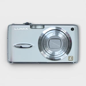 Op de afbeelding: Een zilveren Lumix digitale camera met een Leica lens. De camera heeft een rechthoekige behuizing met een lens aan de voorkant en het woord "LUMIX" gedrukt aan de linkerbovenkant. De lens heeft de woorden "MEGA O.I.S. WIDE" erop.
