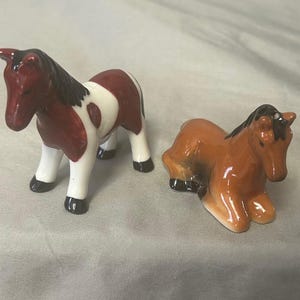 Puede incluir: Dos figuras de caballos de cerámica. Una está de pie, con un cuerpo manchado rojo y blanco, crin negra y patas blancas. La otra está tumbada, con un cuerpo marrón y crin negra. Ambas tienen un acabado brillante.
