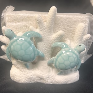May include: A decorative figurine featuring two glossy, light blue sea turtles attached to a textured, white coral-like base. The turtles have a smooth, glazed finish with visible shell patterns, and the entire piece is partially wrapped in clear plastic.