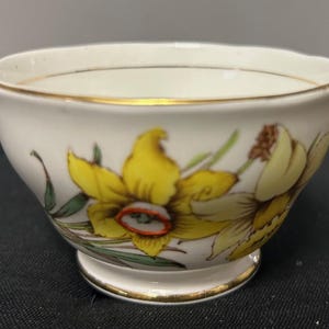 Op de afbeelding: Witte theekop met een gouden rand en voet, versierd met gele narcissenbloemen en groene bladeren. Het bloemmotief bevindt zich aan de buitenkant van de kop. De kop is waarschijnlijk van porselein.
