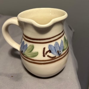May include: A small white ceramic pitcher with a brown and blue floral design. The pitcher has a handle and a wide mouth.