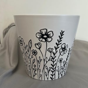 Peut inclure: Pot de fleurs en céramique blanche avec un motif floral noir. Le pot présente une variété de fleurs et de feuillages stylisés, créant une esthétique de jardin. Le design est dessiné à la main, ajoutant une touche unique.