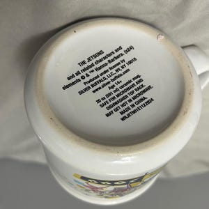 May include: White ceramic mug featuring The Jetsons characters. The mug has a capacity of 20 oz (591 ml) and includes text on the bottom. It is safe for microwave and dishwasher use. Made in China.