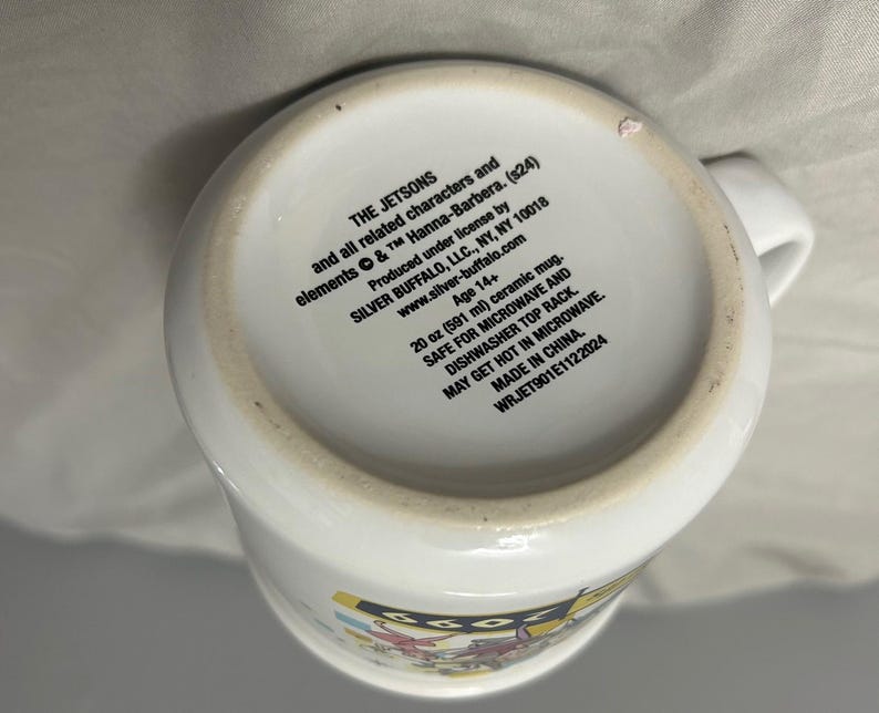 May include: White ceramic mug featuring The Jetsons characters. The mug has a capacity of 20 oz (591 ml) and includes text on the bottom. It is safe for microwave and dishwasher use. Made in China.