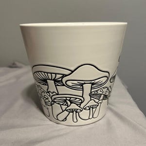 May include: White ceramic planter with a black outline of mushrooms. The planter is a simple, rounded shape with a wide opening.