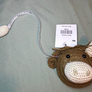 May include: A brown and white crochet horse head tape measure with a white measuring tape extending from the horse's head. The tape measure is labeled with measurements in inches. A tag is attached to the tape measure.