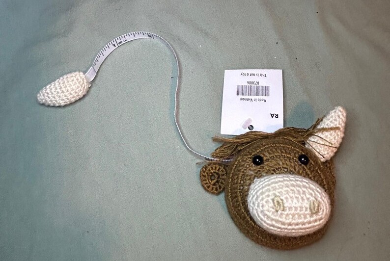 May include: A brown and white crochet horse head tape measure with a white measuring tape extending from the horse's head. The tape measure is labeled with measurements in inches. A tag is attached to the tape measure.