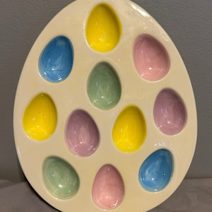 Puede incluir: Un plato de servir de cerámica blanca en forma de huevo con 12 hendiduras individuales en forma de huevo. Las hendiduras están pintadas en colores pastel: amarillo, azul, verde y rosa.