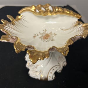 May include: A white porcelain pedestal bowl shaped like a seashell with a scalloped, gold-gilded rim. The interior features a small, central floral decal in pink and green tones. The base is flared and decorated with gold accents and embossed details.