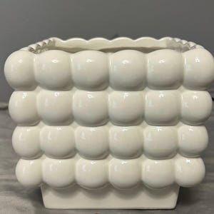 May include: A white ceramic planter with a distinctive bubble-like texture on its exterior. The square planter has a scalloped edge and a rectangular base. The design and white colour give it a modern feel. Suitable for small plants or succulents.
