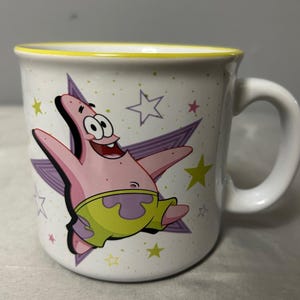 May include: White ceramic mug featuring Patrick Star from Spongebob Squarepants. The mug has a yellow rim and a white handle. Patrick is depicted with a purple starburst background and yellow and purple stars.