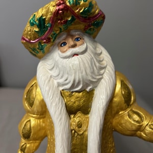 Può includere: Statuina decorativa di Babbo Natale con barba bianca e occhi blu. La figura indossa una veste e un cappello dorati con accenti verdi e rossi. Decorazione festiva per le vacanze.