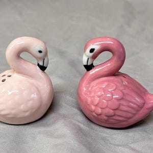 Puede incluir: Dos saleros y pimenteros de cerámica con forma de flamenco. Uno es de color rosa pastel claro y el otro es de un rosa más intenso y vibrante. Ambos tienen cuellos curvos, picos con puntas negras y pequeños orificios en la parte superior.