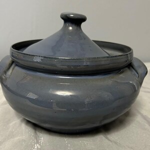 May include: A blue ceramic casserole dish with a lid. The dish has a rounded body with two handles and a lid with a small knob. The glaze has a glossy finish.