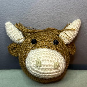 May include: A crocheted cow head with a brown body, white muzzle, and white horns. The cow has black eyes and a fringe of brown yarn on top of its head. The handmade item is likely a toy or decorative piece.