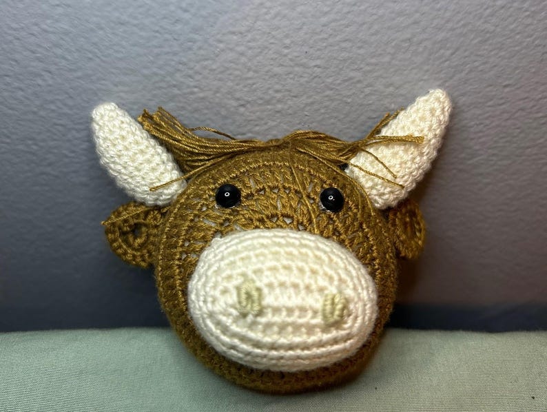 May include: A crocheted cow head with a brown body, white muzzle, and white horns. The cow has black eyes and a fringe of brown yarn on top of its head. The handmade item is likely a toy or decorative piece.