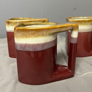 May include: Four ceramic mugs with a distinctive square design and a unique handle. The mugs feature a gradient of colours, starting with a deep red at the base, transitioning to white, and topped with a brown rim. These mugs are ideal for coffee or tea.