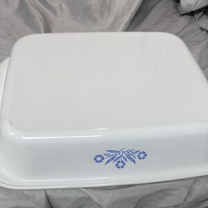 May include: White square baking dish with rounded edges. The dish has a floral design in blue on the side. The dish is made of a glossy material and is sitting on a gray surface.