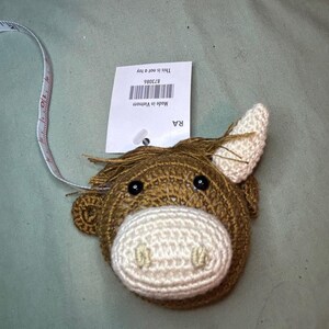 May include: A brown and cream-colored crochet tape measure shaped like a horse's head. The tape measure is white with black markings. A tag attached to the tape measure reads "This is not a toy" and "Made in Vietnam".